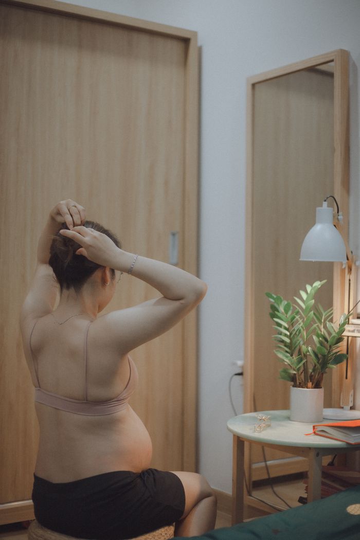 A pregnant woman sits indoors tying her hair in front of a mirror.