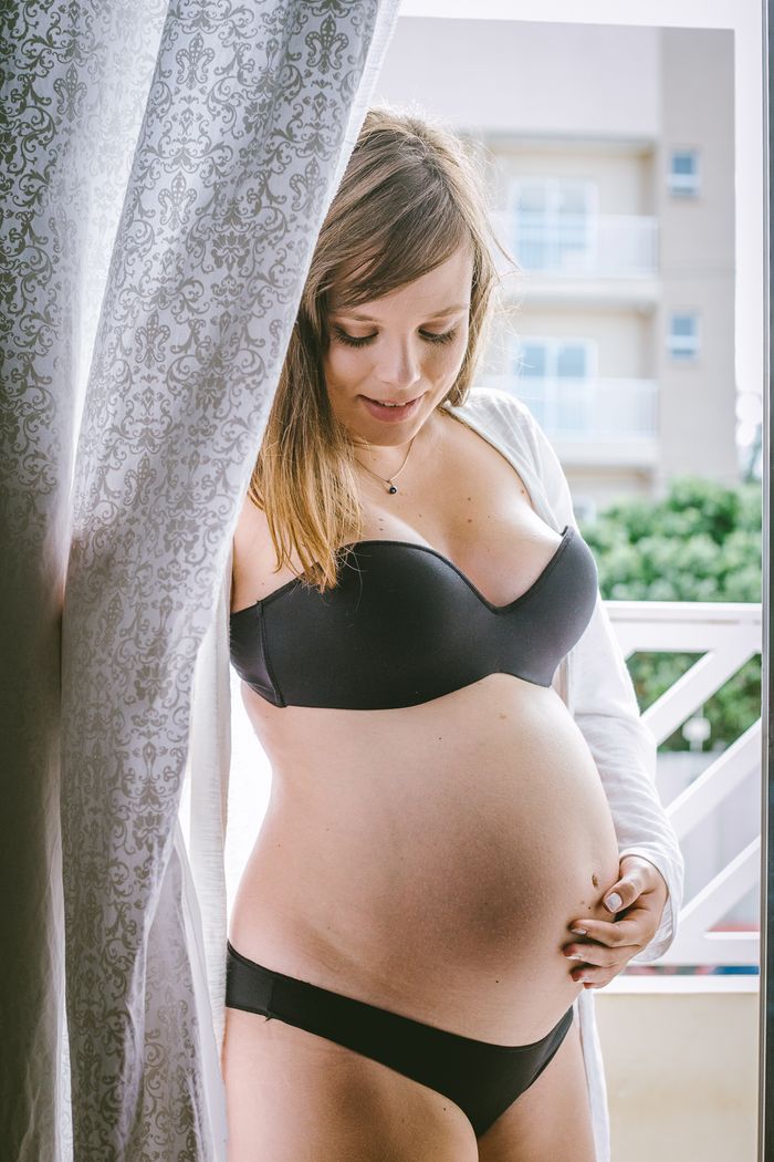 Smiling pregnant woman in black underwear standing on balcony with hand on belly and enjoying pregnancy
