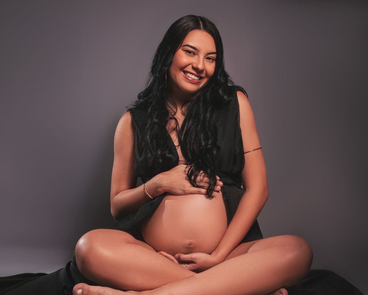 Beautiful pregnant woman smiling, seated in a studio with a gray background.